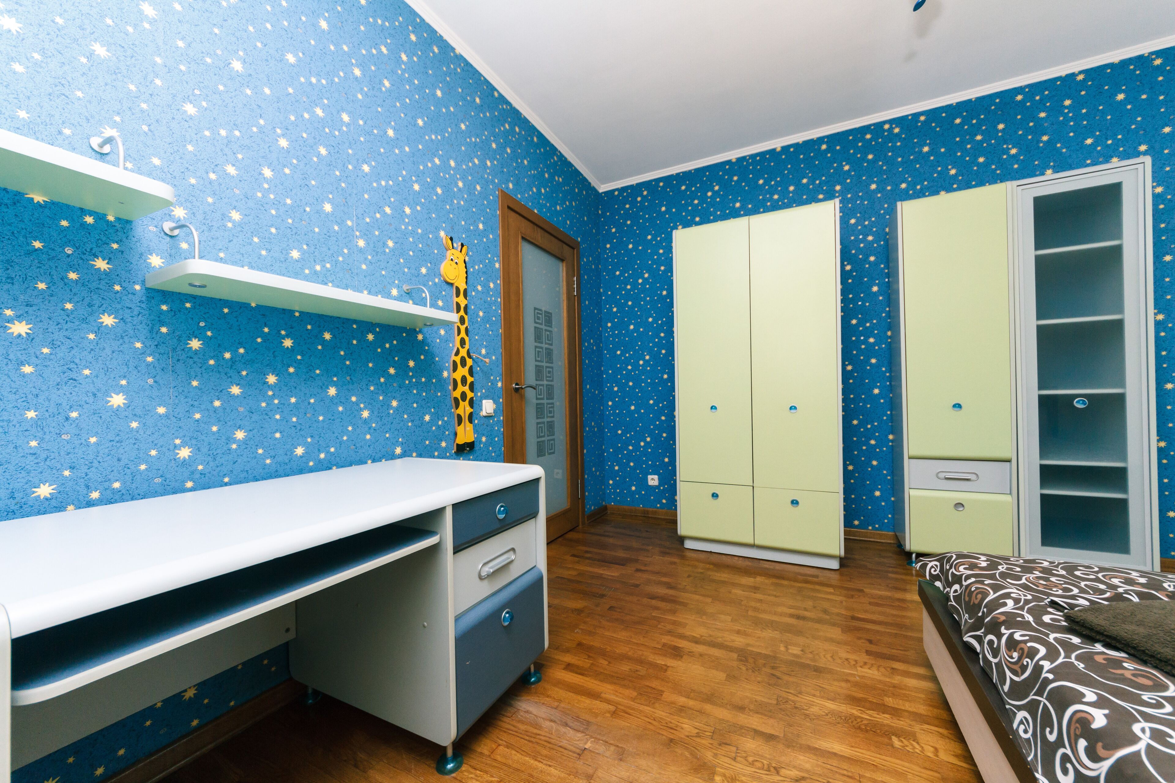 A bedroom with a star-patterned wall, a desk with drawers, a bed with a patterned bedspread, and two wardrobes.