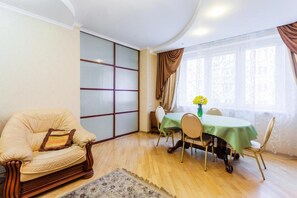 Une pièce avec un plancher en bois, un fauteuil beige avec un coussin, une table à manger recouverte d’une nappe verte et une fenêtre avec des rideaux.