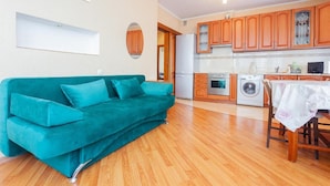A compact living space with a teal sofa, wooden flooring, a kitchen with wooden cabinets, a refrigerator, a washing machine, and a dining table with a tablecloth. - Comfortable apartment on the left bank (Kyiv)