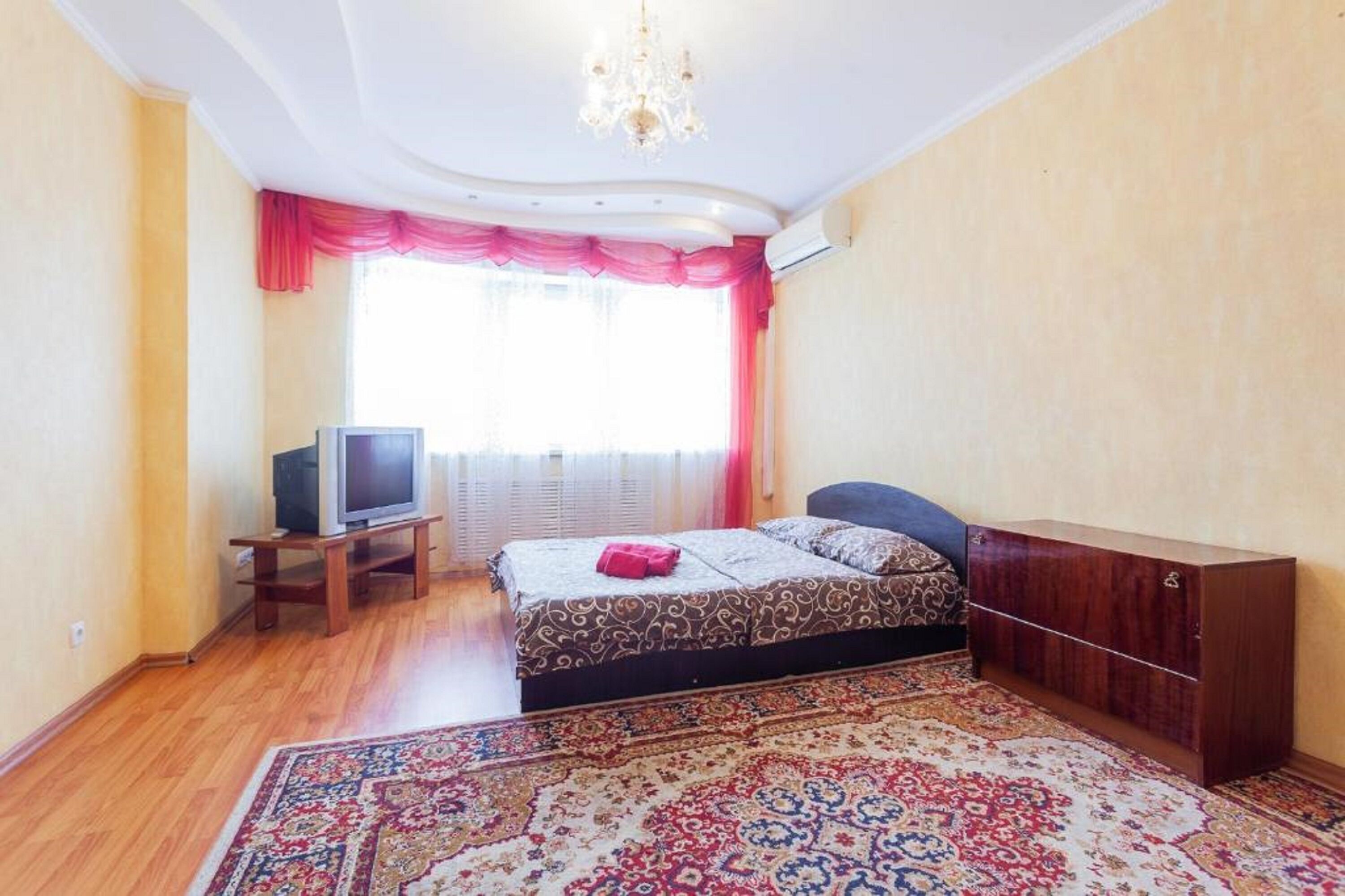 A bedroom with a bed, a television on a stand, a wooden wardrobe, and a patterned rug.