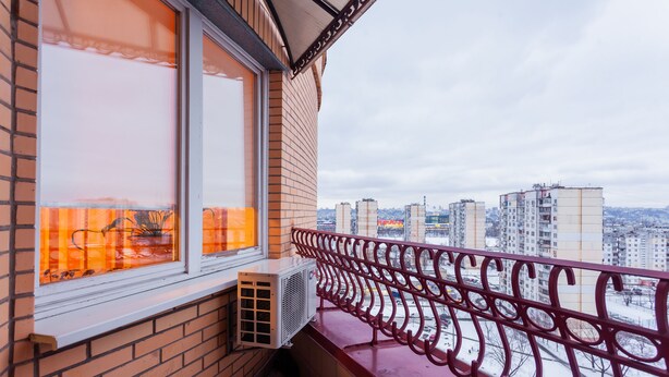 Un balcon avec une balustrade rouge, une fenêtre donnant sur de hauts immeubles et un mur de briques.