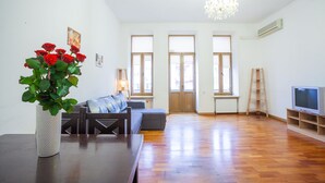 Een ruime woonkamer met houten vloer, een grijze bank, een witte vaas met rode bloemen en een televisie op een houten dressoir.