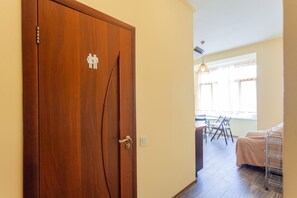 Uma porta de madeira com símbolos de banheiro masculino e feminino, uma área de refeições com cadeiras e uma mesa, e uma cama com estrutura de metal.