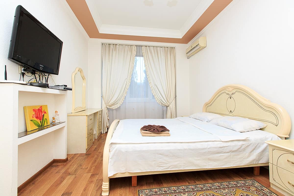 A bedroom with a bed, a television on a stand, a dresser, a mirror, and a window with curtains.