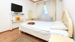 Un dormitorio con una cama, televisión en un estante, mesita de noche, espejo y ventana con cortinas.