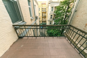 A balcony with a tiled floor and metal railing, overlooking a narrow street with buildings and trees.
