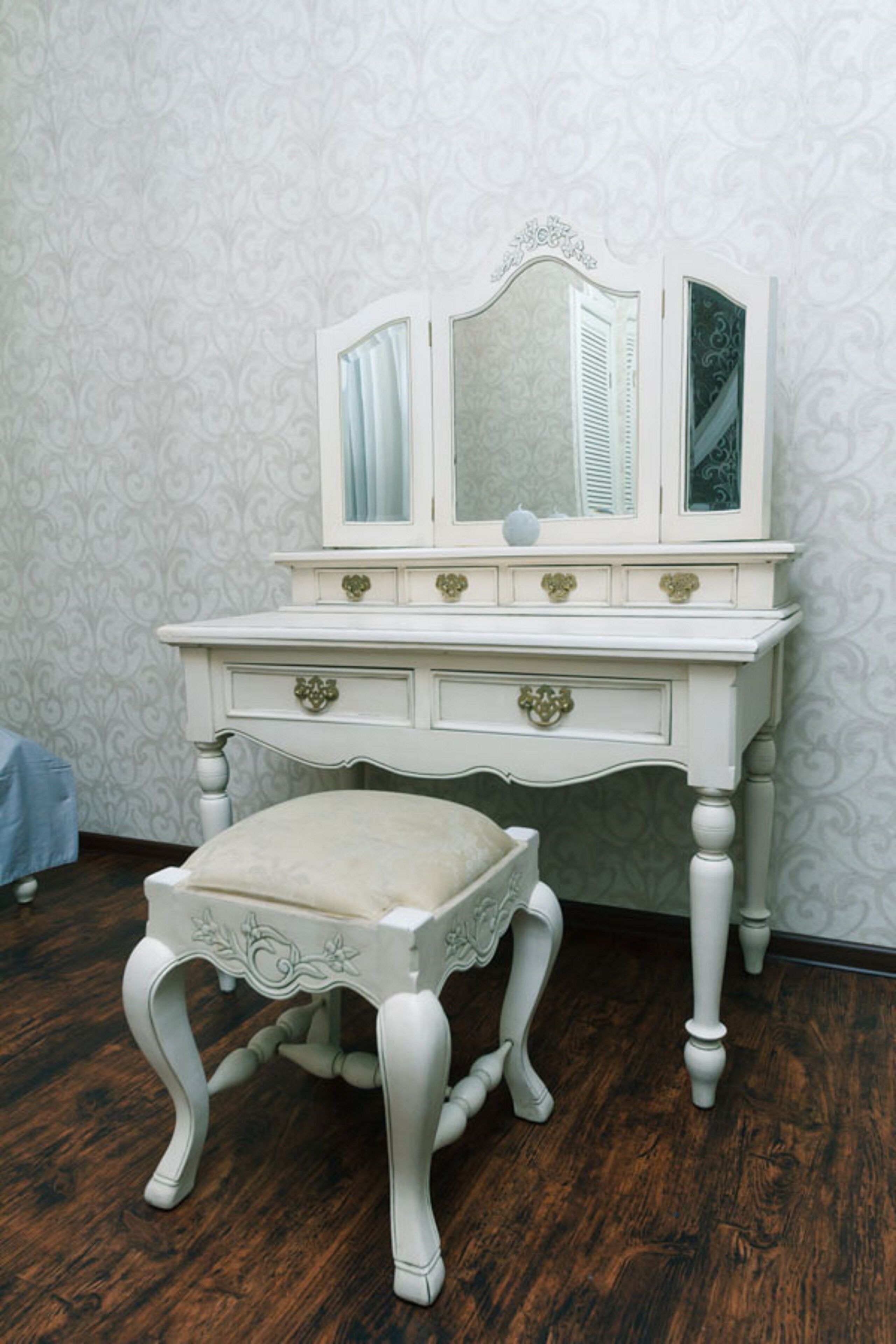 A white vanity with a mirror, drawers, and a cushioned stool.