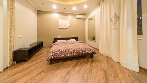 A bedroom with a wooden floor, a bed with a patterned bedspread, a bedside table, a mirror, and a wall-mounted air conditioner.