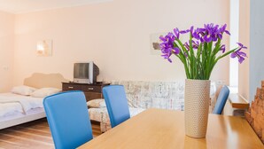 Una habitación con una mesa de madera, sillas azules, una cama con ropa de cama blanca, un televisor sobre un mueble bajo y un jarrón con flores moradas.