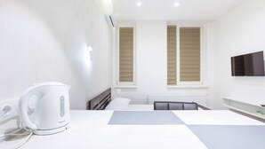 Ein modernes Hotelzimmer mit einem weißen Bett, einem flachbildfernseher an der Wand, einem weißen Wasserkocher auf dem Nachttisch und einem Fenster mit Jalousien.