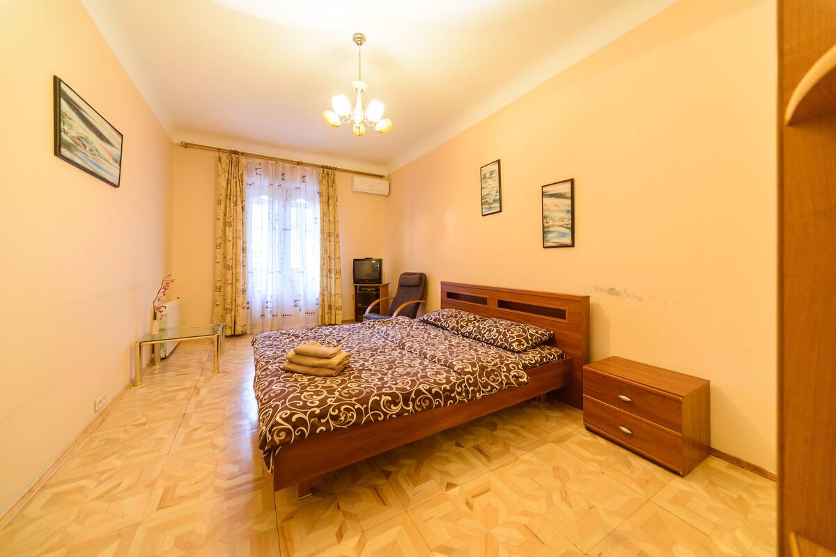 A bedroom with a large bed, a wooden nightstand, a chair, and a ceiling light.