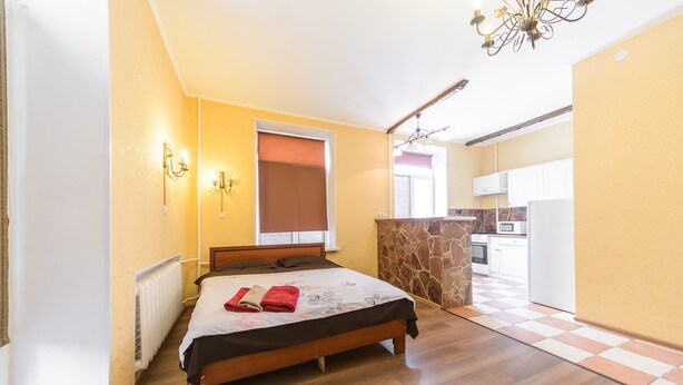 A bedroom with a wooden bed, a radiator, a window with a blind, and a kitchen in the background. - Cozy studio apartment near Palace Ukraina (Kyiv)