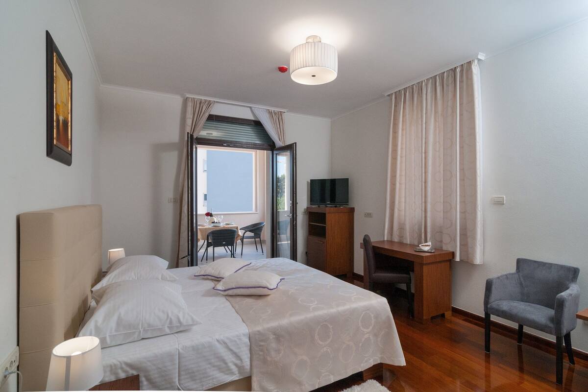A bedroom with a bed, bedside lamps, a desk, a chair, a television, and a window with curtains.