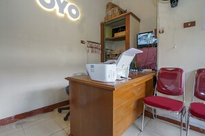 Een receptiebalie met een computer, telefoon en monitor. Er zijn twee rode stoelen en een houten kast met dozen erop.