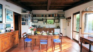 A cozy kitchen with a wooden table, chairs, and a tiled floor. There are bookshelves, a refrigerator, and a window providing natural light.