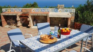 An outdoor dining area with a tiled table and chairs, a brick oven, and a view of the sea.