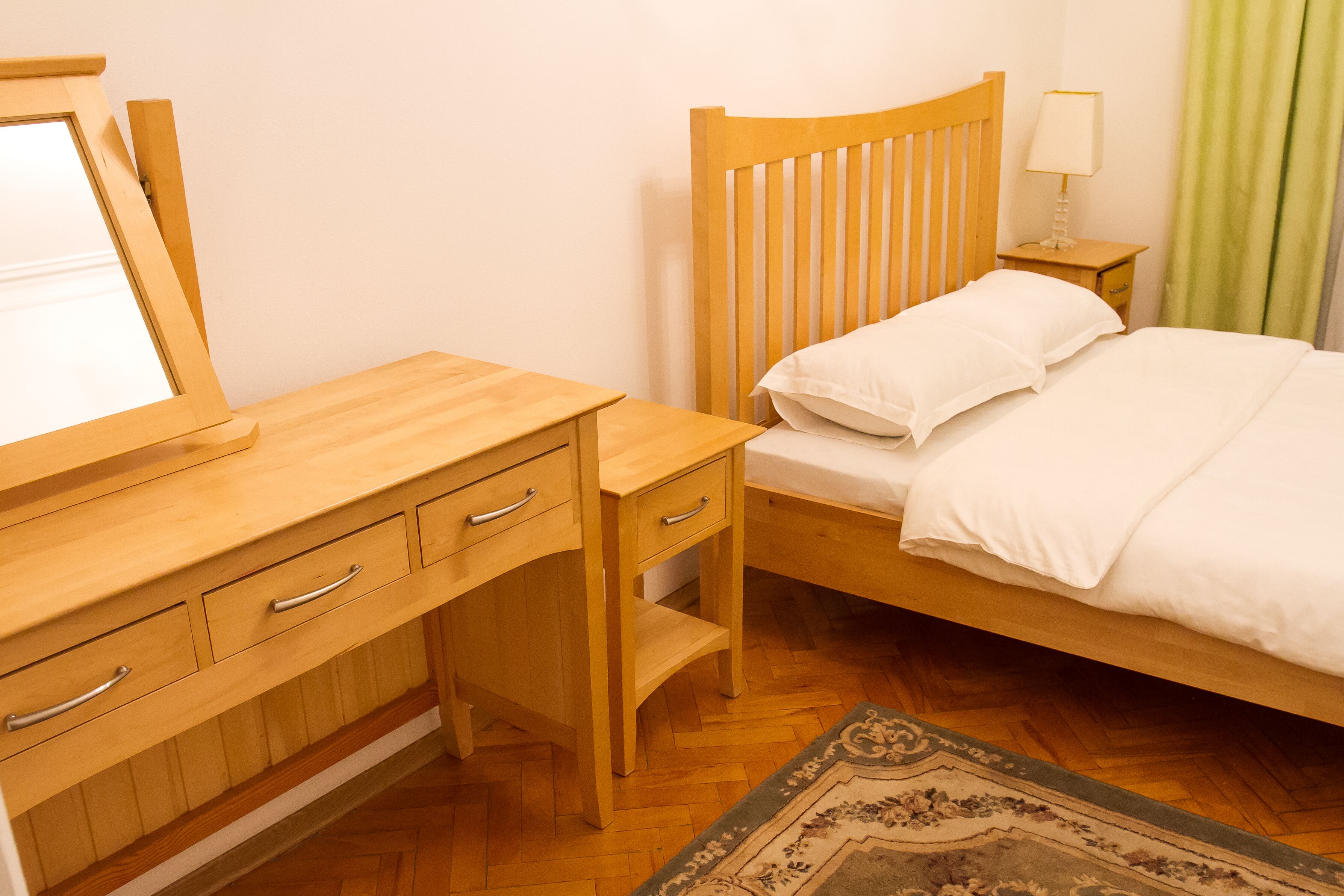 Una cama de madera con ropa de cama blanca, un escritorio de madera con cajones, una mesita de noche con una lámpara y una alfombra estampada sobre un piso de madera.