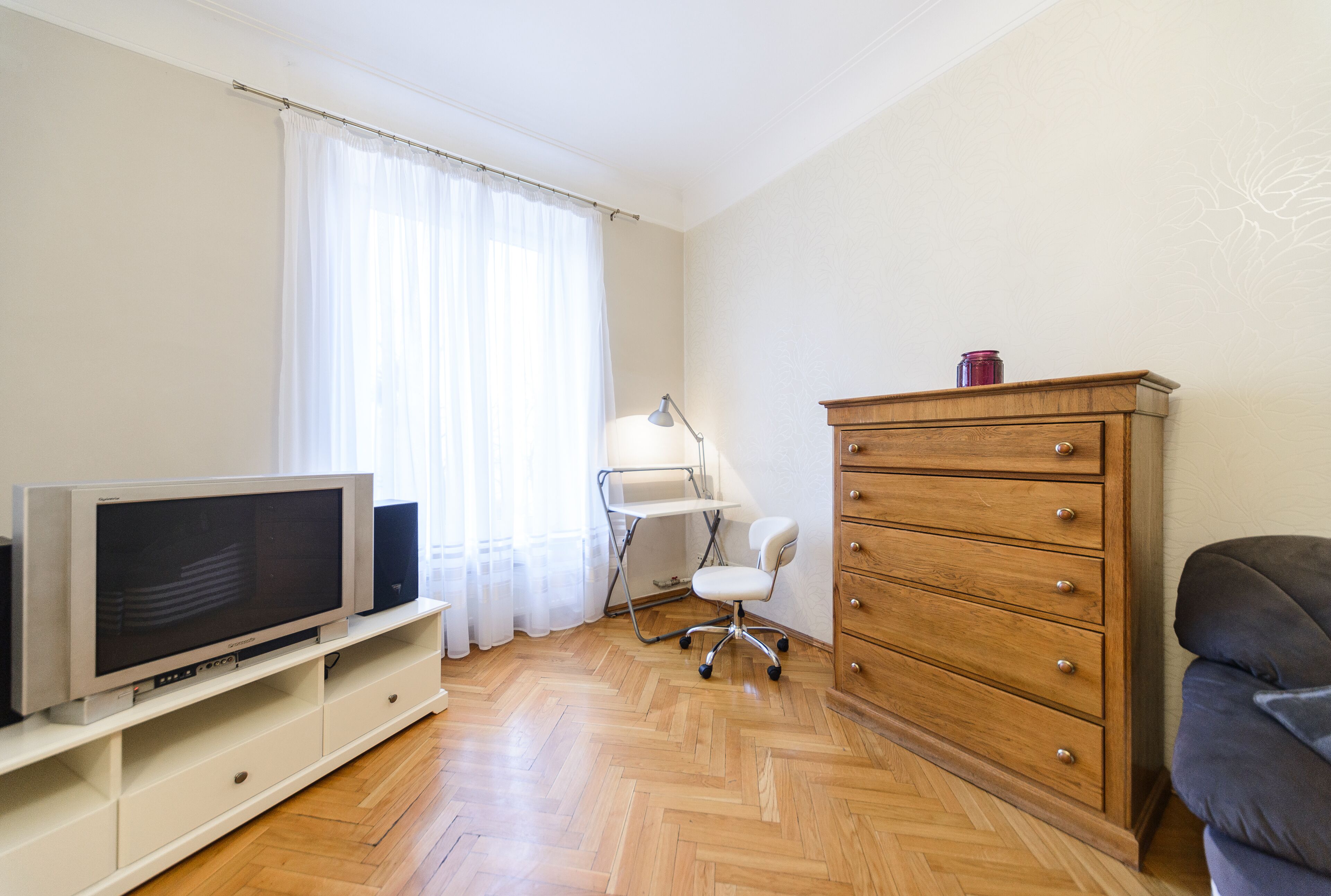 Une pièce comprenant une télévision posée sur un meuble blanc, une commode en bois, un bureau avec une lampe et une chaise.