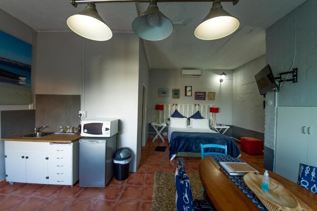 A compact room with a kitchenette, a bed with a blue bedspread, a small table with a chair, and a television mounted on the wall.