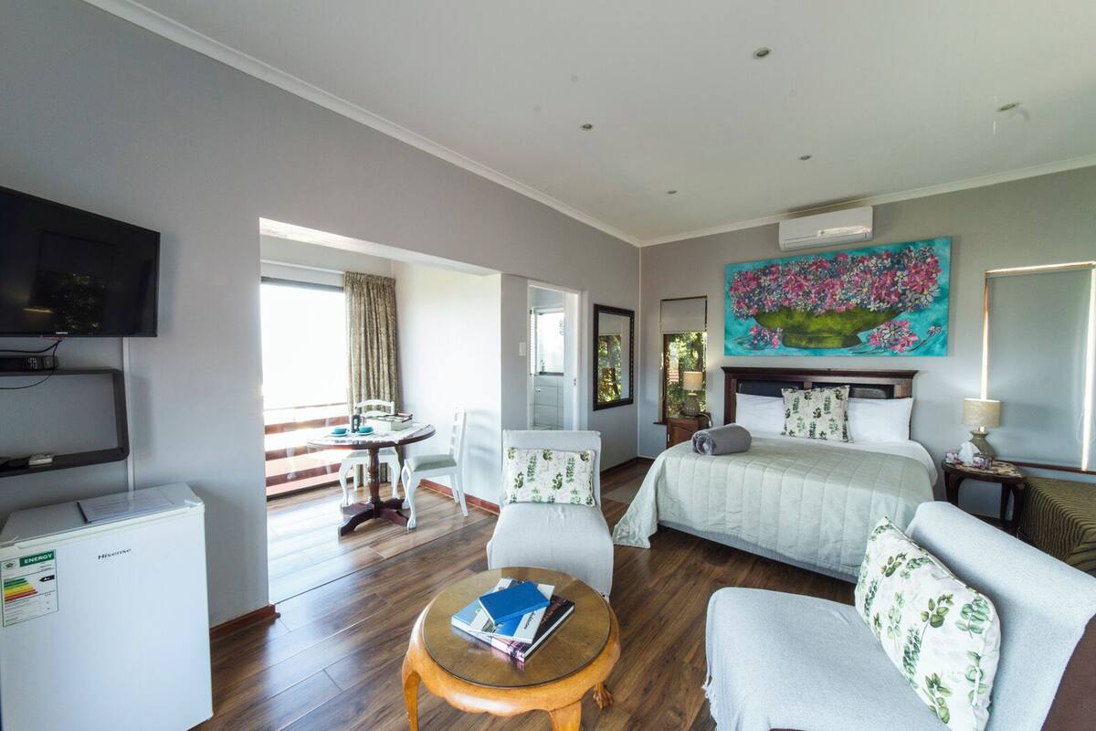 A hotel room with a bed, a TV, a desk, and a chair. There is a painting on the wall and a window with curtains.