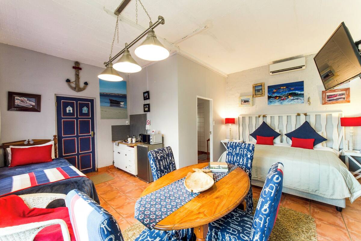 A cozy room with a bed, a dining table with chairs, a small kitchen area, and a door with a nautical theme.