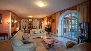 A spacious living room with a large sofa, a coffee table, and a television. There are arched windows and a door leading to another room. - Villa Amalia Resort (Cava de' Tirreni)