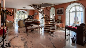 A spacious living room with a grand piano, a staircase, and a large window with a view of a building. - Villa Amalia Resort (Cava de' Tirreni)