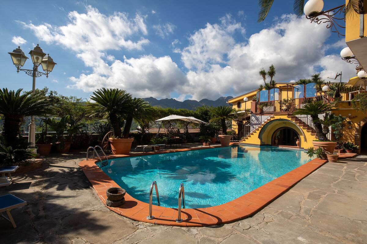 Uma área de piscina com piscina azul cristalina, piso de pedra e vista para montanhas ao fundo.