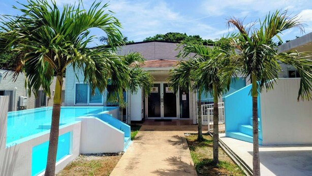A pool area with palm trees, a building entrance, and a clear sky.