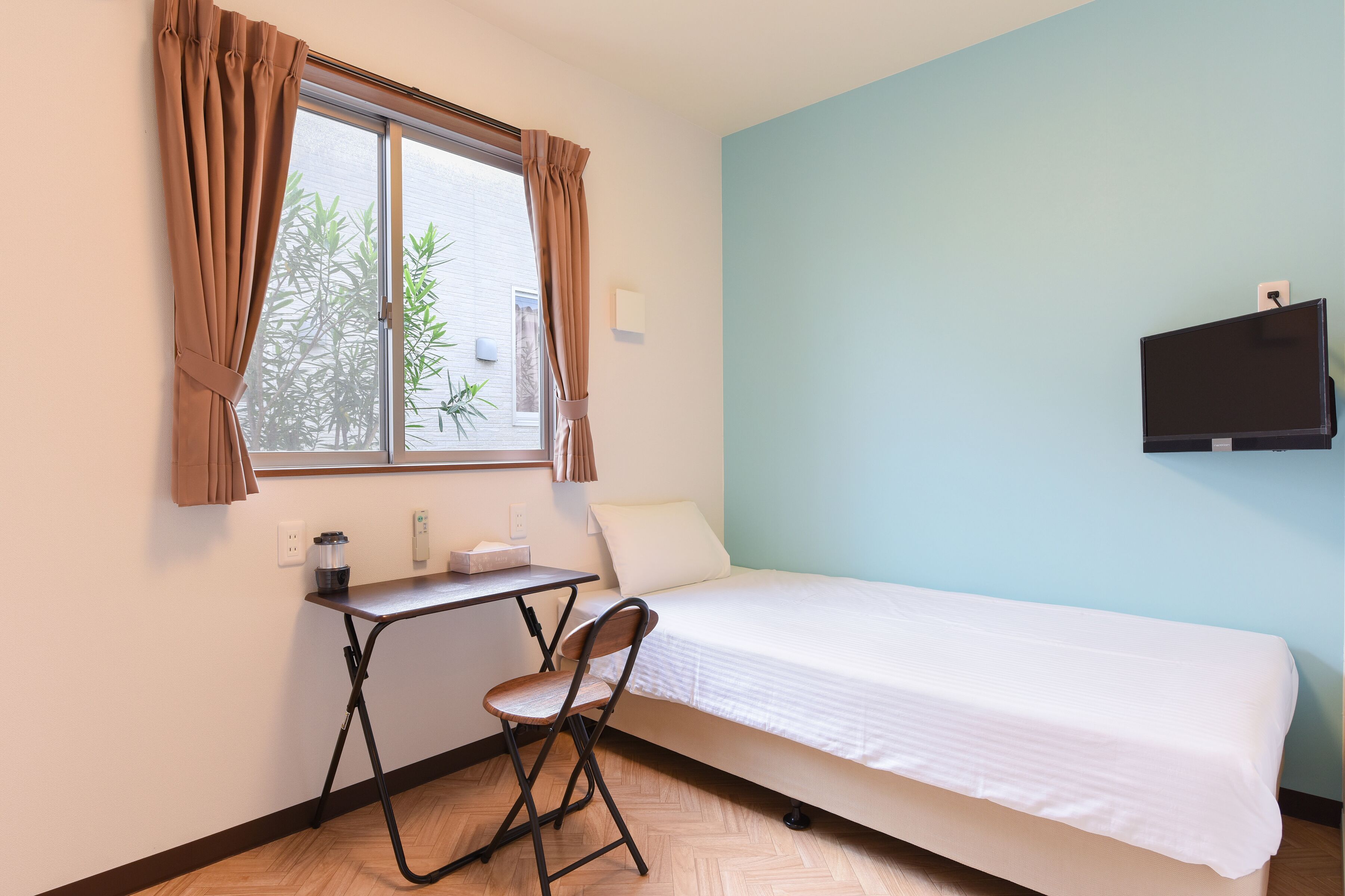Een kleine kamer met een bed, een bureau, een stoel, een raam met gordijnen en een aan de muur bevestigde televisie.