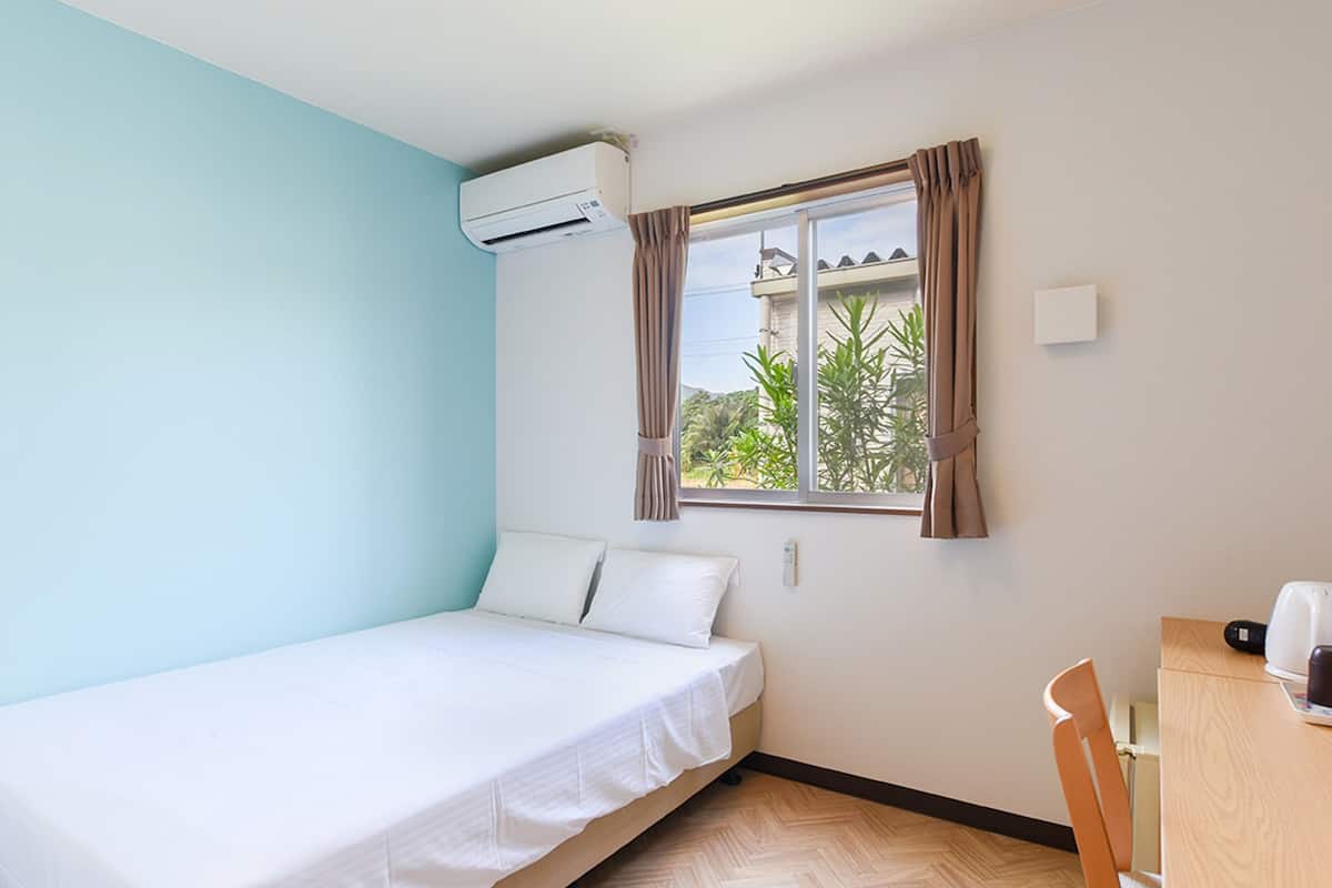 A small, single-bed room with a window, a desk, and an air conditioning unit.