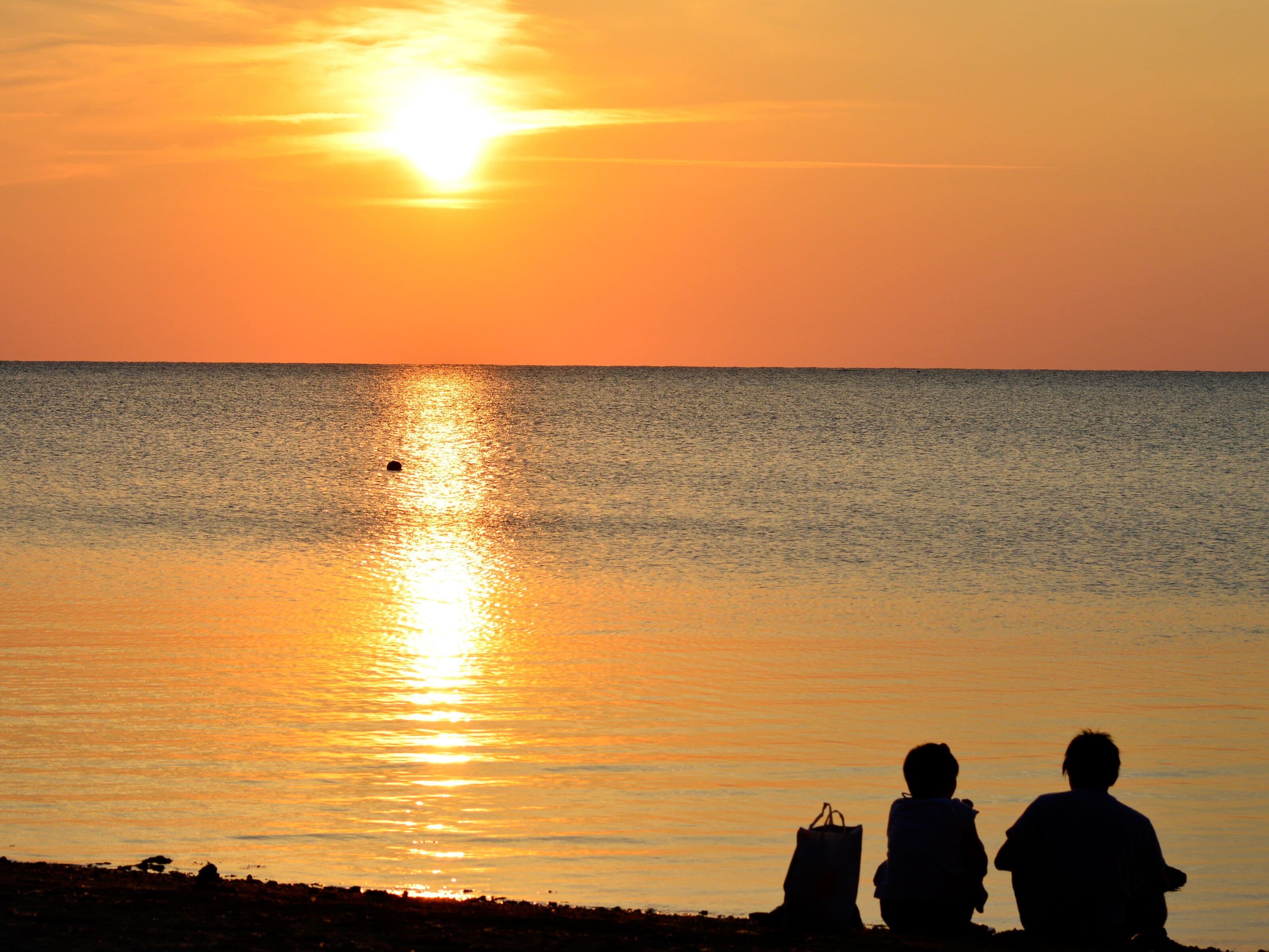 A sunset over a calm sea with silhouettes of two people and a bag on the shore.