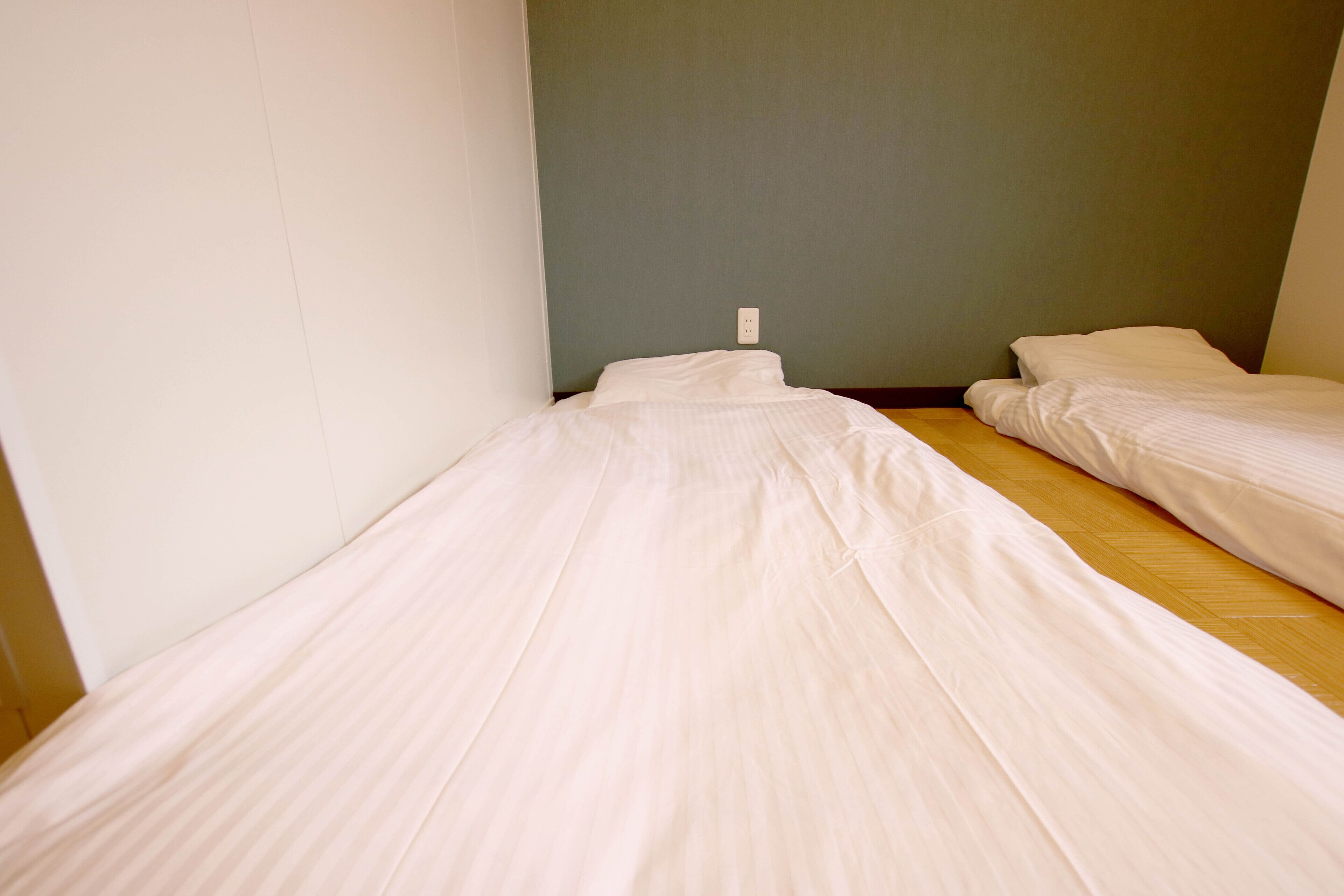 A small, single-room with two beds, a wooden floor, and a plain wall.