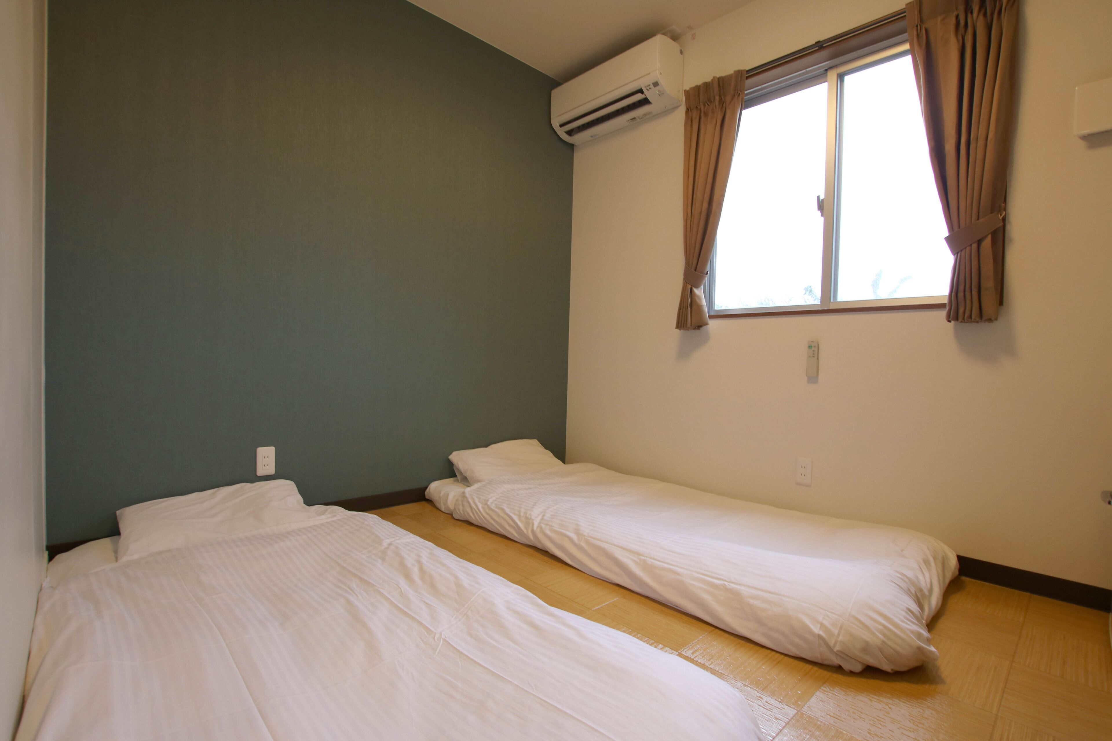 A room with two single beds, a window with curtains, an air conditioning unit, and a light switch on the wall.