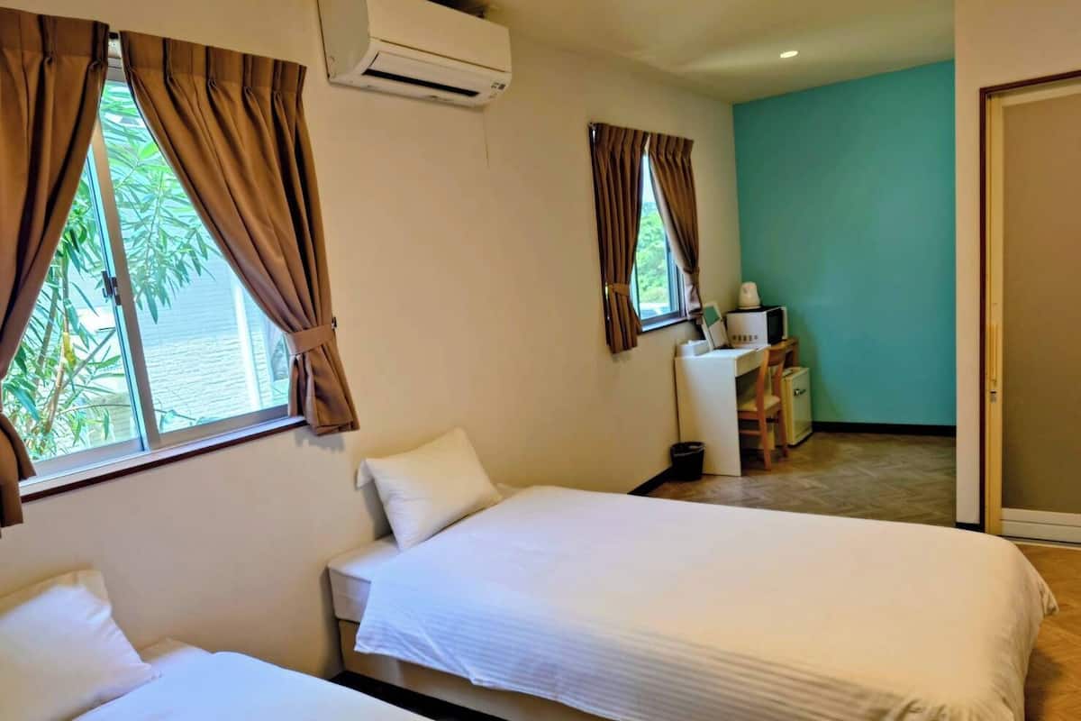 A hotel room with two beds, a desk with a computer, an air conditioner, and a view of greenery outside.