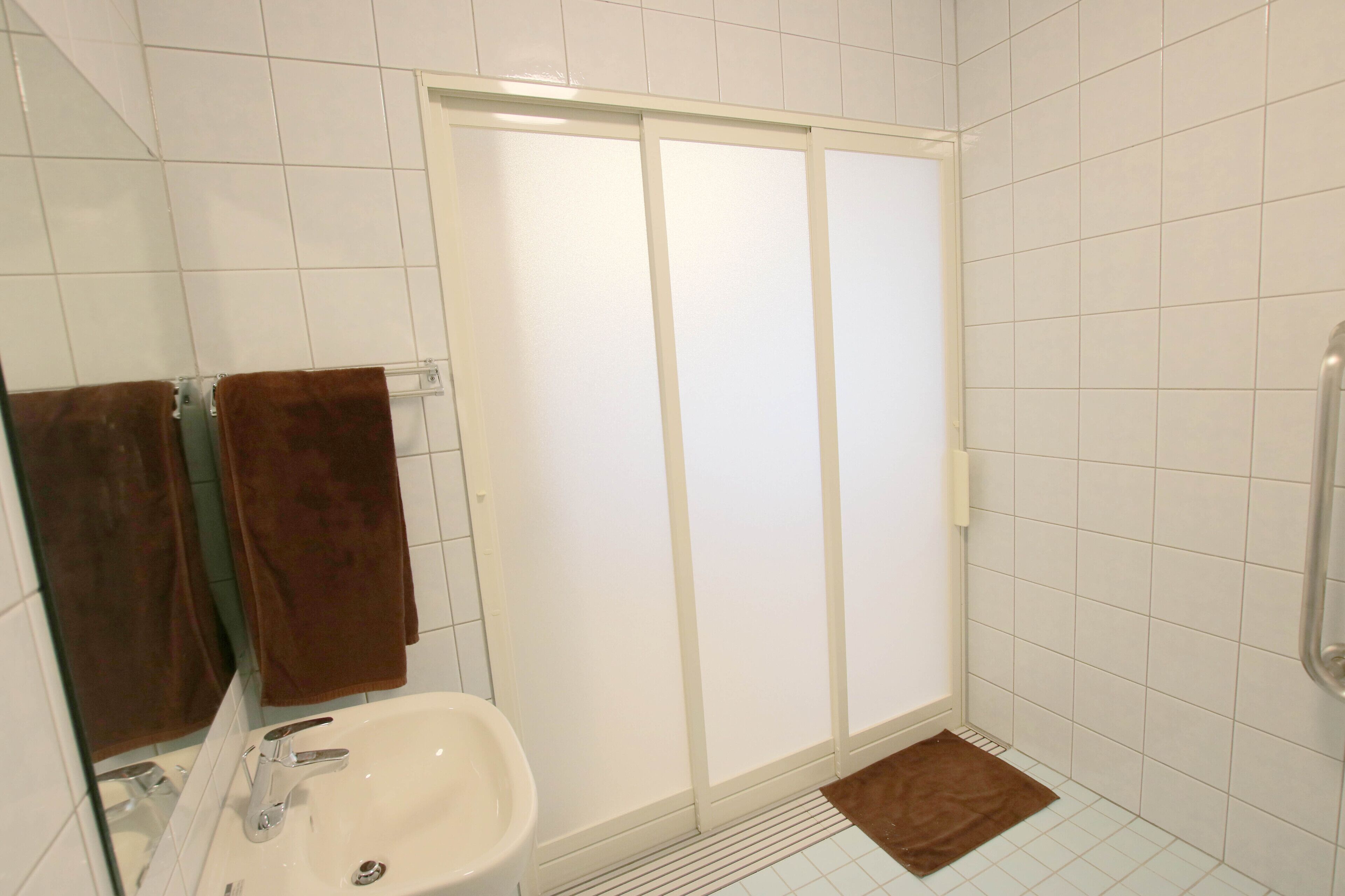 Une salle de bain avec un lavabo blanc, un miroir, un porte-serviettes avec des serviettes marron, une douche avec une porte en verre dépoli et un sol carrelé blanc.