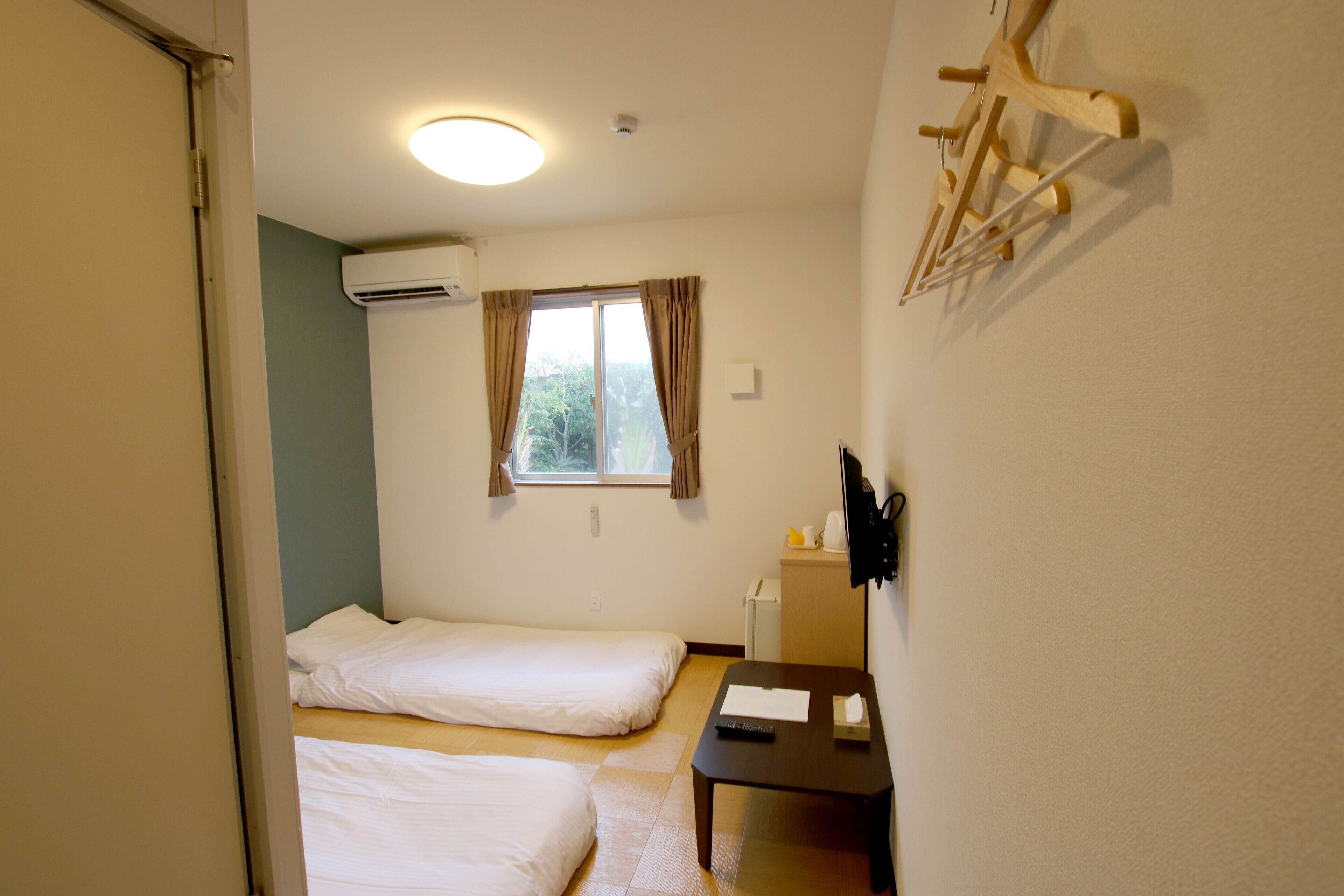 Una habitación pequeña y limpia con dos camas individuales, una mesa de madera con un control remoto, un mueble pequeño y una ventana con cortinas.