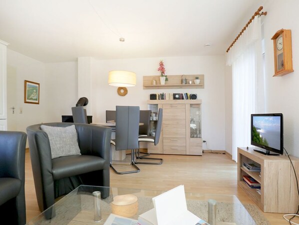A modern living room with a glass coffee table, a sofa, a TV, and a dining area with chairs and a kitchen in the background. - Vacationer village 20c, Germany (Ostseebad Boltenhagen)