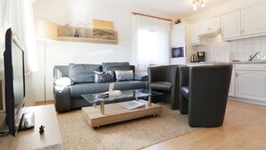 Un soggiorno moderno con un divano grigio, un tavolino in vetro e una TV. È presente un angolo cottura con pensili bianchi e un forno a microonde.