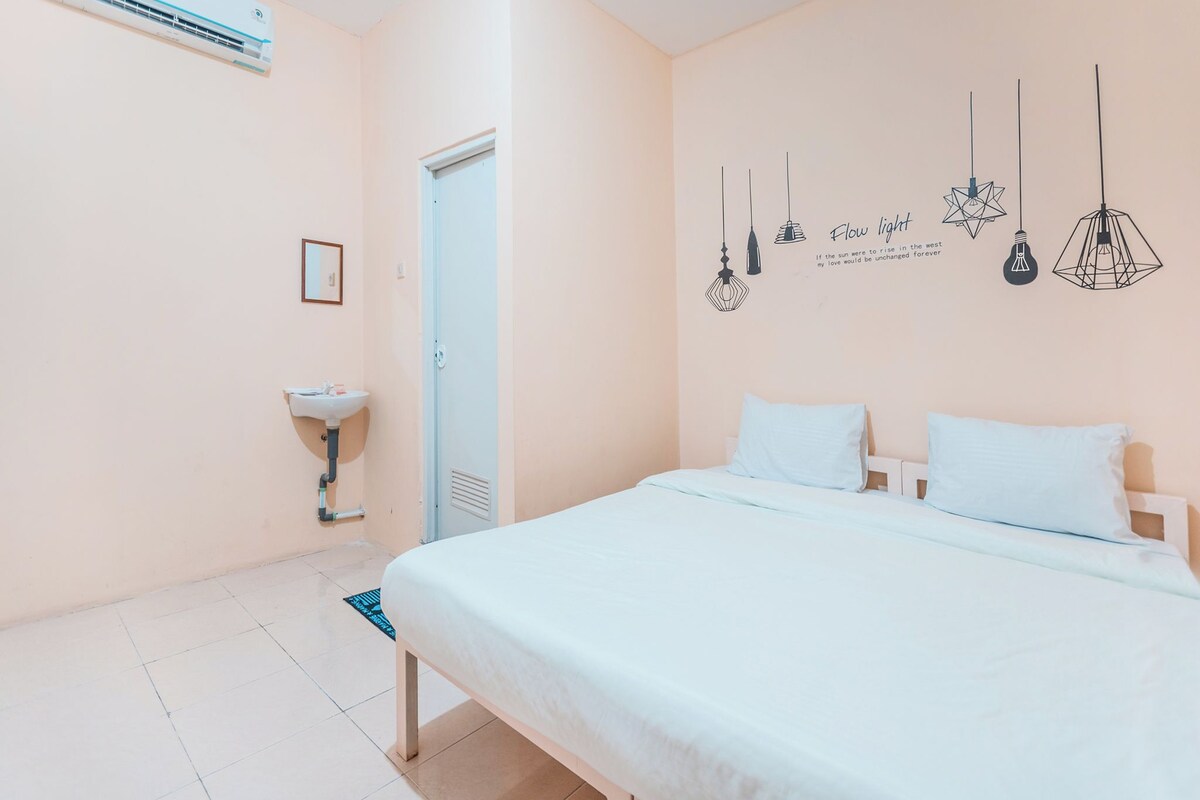 Une chambre à coucher comprenant un lit, une table de chevet, un climatiseur fixé au mur, un luminaire fixé au mur et une porte.