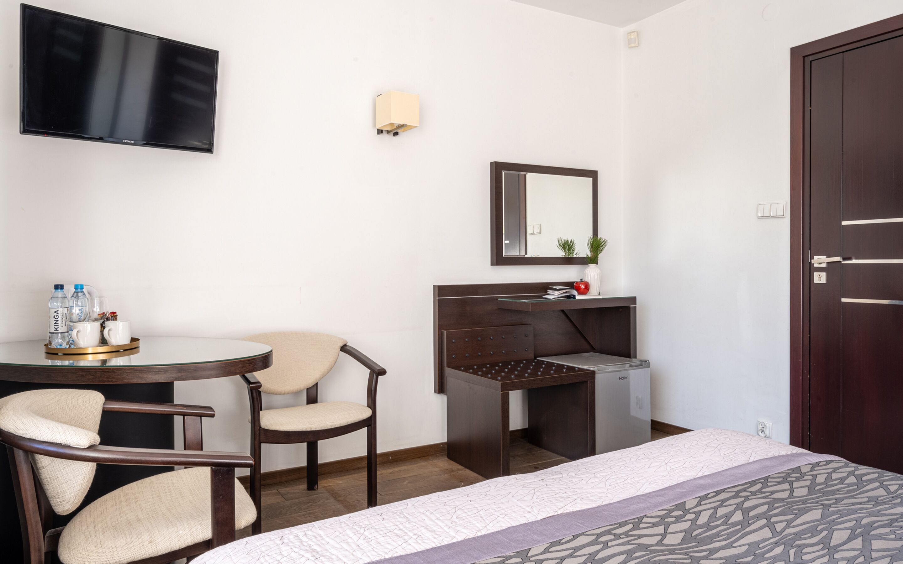 A hotel room with a bed, a desk with a mirror, a TV mounted on the wall, and a chair.
