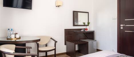 A hotel room with a bed, a desk with a mirror, a TV mounted on the wall, and a chair.