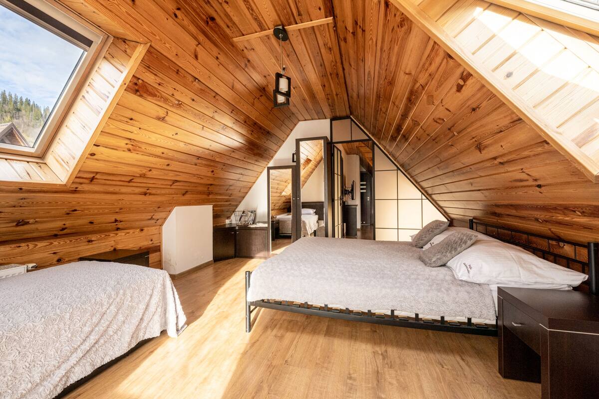 A cozy attic bedroom with wooden walls and sloped ceilings, featuring two beds and a skylight.