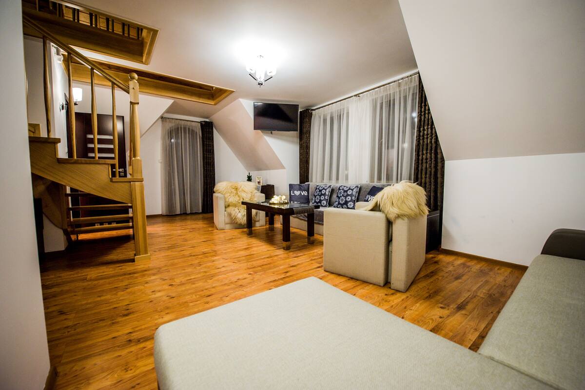 A living room with a wooden staircase, a sofa set, a coffee table, and a flat-screen TV mounted on the wall.