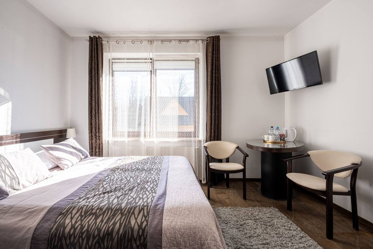 A hotel room with a bed, two chairs, a small table with a tray, a TV mounted on the wall, and a window with curtains.