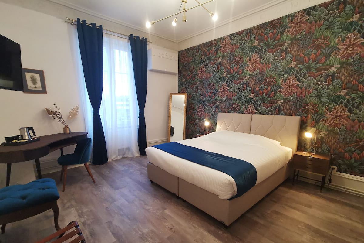 A bedroom with a bed, a desk, a chair, a mirror, and a wall with a floral pattern.