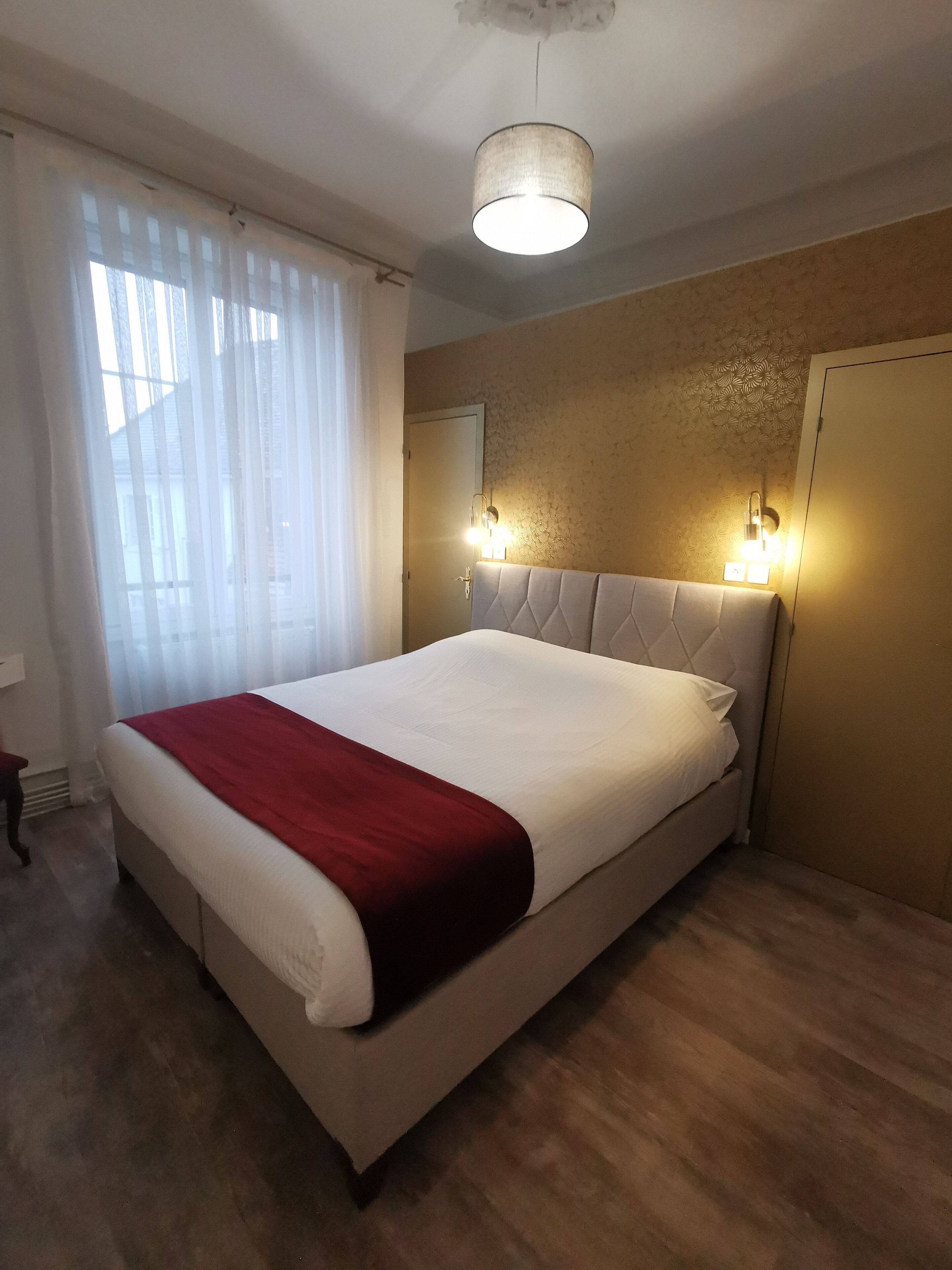 Un grand lit recouvert d’un plaid rouge, un luminaire suspendu et deux lampes fixées au mur.