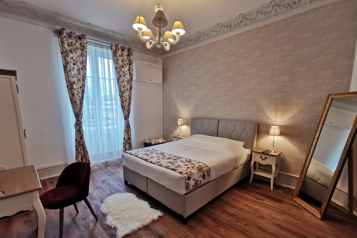 A bedroom with a bed, two bedside tables with lamps, a chair, a mirror, and a window with curtains.