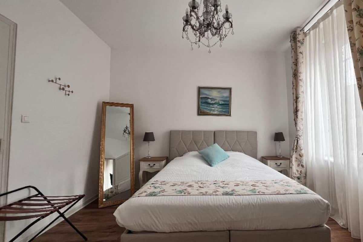 A bedroom with a large bed, a chandelier, a mirror, and a window with curtains.