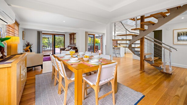 Uma ampla área de jantar com uma mesa de madeira posta para uma refeição, uma escada que leva a um andar superior e uma vista do exterior através de grandes janelas.